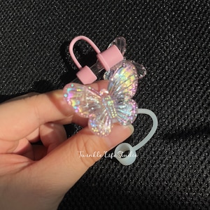 May include: A clear glass bottle stopper with a pink silicone ring and a iridescent butterfly charm. The stopper is attached to a white silicone strap.