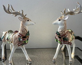 May include: Two decorative deer figurines. The deer have white bodies, tan fur, and gold-colored antlers. They are adorned with festive red, green, and gold accents around their necks and backs, suggesting a holiday theme. The figurines are displayed on a reflective surface.