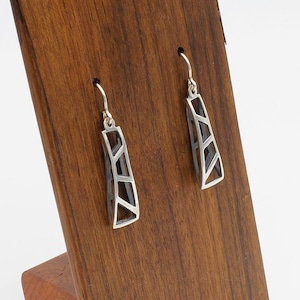 May include: A pair of silver dangle earrings with a geometric design. The earrings are hanging on a wooden display stand.
