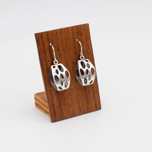May include: A pair of silver earrings with a geometric, openwork design. The earrings are displayed on a wooden stand.