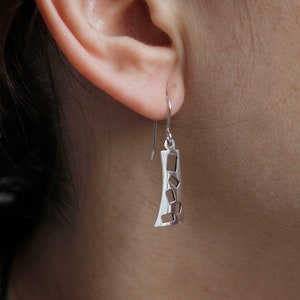 Puede incluir: Pendientes de plata con diseño geométrico. Los pendientes están hechos de una fina pieza rectangular de metal con recortes en forma de cuadrados. Los pendientes cuelgan de un simple gancho.