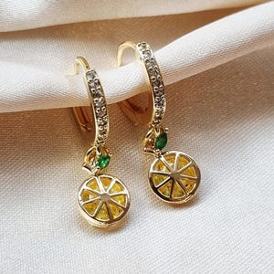 Puede incluir: Un par de pendientes de aro dorados. Cada pendiente presenta un pequeño dije redondo que se asemeja a una rodaja de limón con detalles amarillos y verdes. Los aros están adornados con pequeños cristales transparentes.