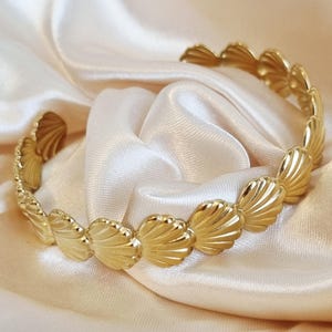 May include: A gold-toned cuff bracelet featuring a repeating pattern of small, fan-shaped seashell motifs. The bracelet has an open design, allowing it to be slipped on the wrist, and displays a polished, reflective metallic finish throughout.