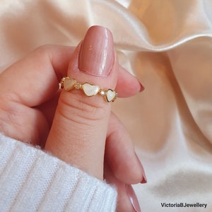 Peut inclure: Bague en or avec une chaîne délicate et cinq charmes en forme de cœur blancs. Chaque charme a un petit strass transparent au centre.