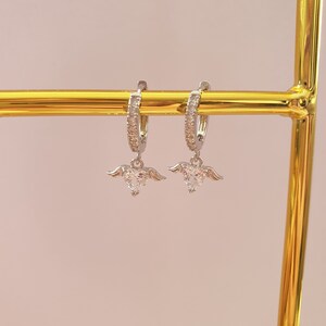 Puede incluir: Un par de pendientes de aro de plata con un peque&ntilde;o dije de coraz&oacute;n de cristal transparente que cuelga de cada aro. Los dijes de coraz&oacute;n tienen peque&ntilde;as alas plateadas a cada lado.