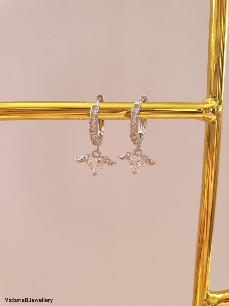 Puede incluir: Un par de pendientes de aro de plata con un peque&ntilde;o dije de coraz&oacute;n de cristal transparente que cuelga de cada aro. Los dijes de coraz&oacute;n tienen peque&ntilde;as alas plateadas a cada lado.