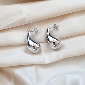 May include: A pair of silver teardrop-shaped earrings. The earrings are made of a smooth, shiny metal and have a simple, modern design.