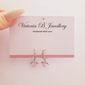 Puede incluir: Aretes de aro de plata con un dije de coraz&oacute;n con alas. Los aretes est&aacute;n adornados con peque&ntilde;as piedras de circonita c&uacute;bica transparentes. Los aretes se muestran en una tarjeta blanca con el texto "Victoria B Jewellery Handmade With Love".