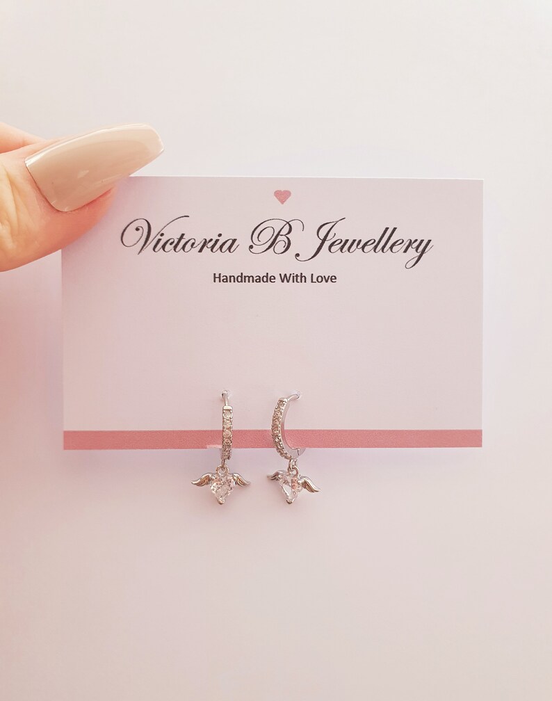 Puede incluir: Aretes de aro de plata con un dije de coraz&oacute;n con alas. Los aretes est&aacute;n adornados con peque&ntilde;as piedras de circonita c&uacute;bica transparentes. Los aretes se muestran en una tarjeta blanca con el texto "Victoria B Jewellery Handmade With Love".
