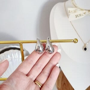 K&ouml;nnte beinhalten: Ein Paar silberne Ohrringe in Tropfenform. Die Ohrringe befinden sich auf einem goldenen St&auml;nder.