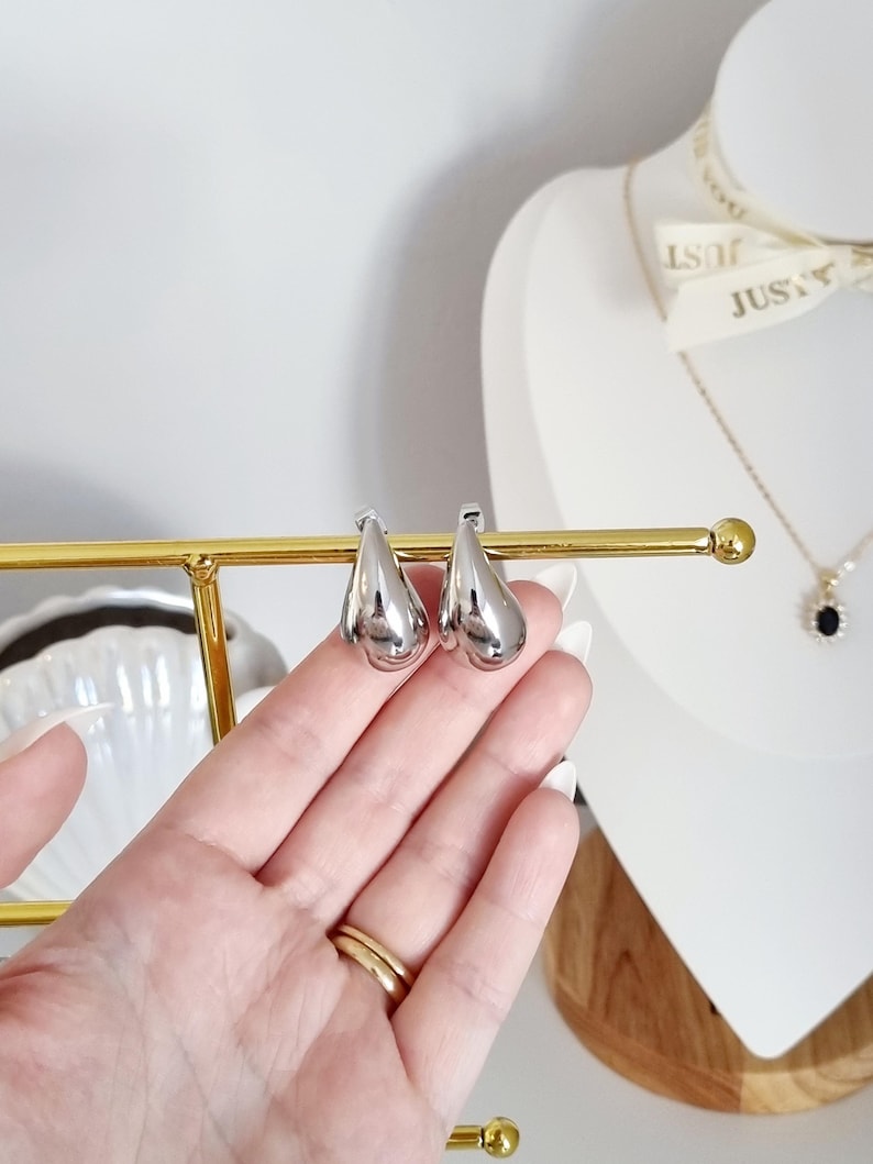 K&ouml;nnte beinhalten: Ein Paar silberne Ohrringe in Tropfenform. Die Ohrringe befinden sich auf einem goldenen St&auml;nder.