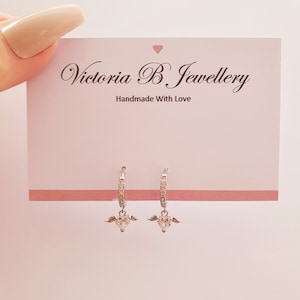 Puede incluir: Aretes de aro de plata con un dije de coraz&oacute;n colgante adornado con piedras de circonita c&uacute;bica transparentes y peque&ntilde;as alas de &aacute;ngel. Los aretes se muestran en una tarjeta rosa con el texto "Victoria B Jewellery Handmade With Love".