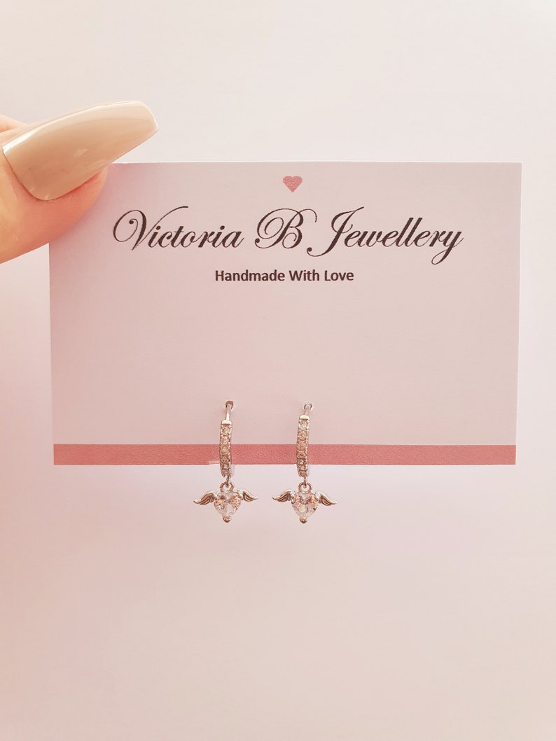 Puede incluir: Aretes de aro de plata con un dije de coraz&oacute;n colgante adornado con piedras de circonita c&uacute;bica transparentes y peque&ntilde;as alas de &aacute;ngel. Los aretes se muestran en una tarjeta rosa con el texto "Victoria B Jewellery Handmade With Love".