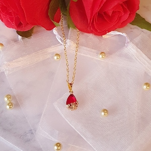 Könnte beinhalten: Eine goldene Kettenkette mit einem roten tropfenförmigen Anhänger. Der Anhänger ist von einem goldenen Filigranmuster umgeben. Die Kette wird auf einem weißen Hintergrund mit roten Rosen und weißem Tüll präsentiert.