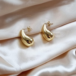 May include: A pair of gold-toned teardrop-shaped earrings. The earrings are made of a smooth, shiny metal and have a simple, modern design.