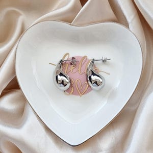 K&ouml;nnte beinhalten: Ein Paar silberne, tropfenf&ouml;rmige Ohrringe auf einer wei&szlig;en herzf&ouml;rmigen Schale mit dem Wort "hello" und einem rosa Herz darauf.