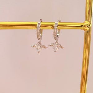 Puede incluir: Un par de pendientes de aro de plata con un peque&ntilde;o dije de coraz&oacute;n de cristal transparente que cuelga de cada aro. Los aros est&aacute;n adornados con peque&ntilde;os cristales transparentes.