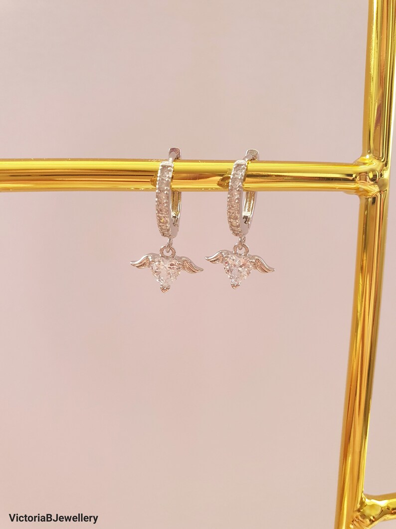 Puede incluir: Un par de pendientes de aro de plata con un peque&ntilde;o dije de coraz&oacute;n de cristal transparente que cuelga de cada aro. Los aros est&aacute;n adornados con peque&ntilde;os cristales transparentes.