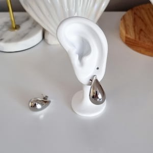 K&ouml;nnte beinhalten: Ein Paar silberne Ohrringe in Tropfenform. Die Ohrringe befinden sich auf einem wei&szlig;en Pr&auml;sentationsst&auml;nder und einer wei&szlig;en Oberfl&auml;che.