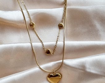 May include: A gold layered necklace with a large heart pendant and smaller heart charms. The necklace is on a light beige fabric background.