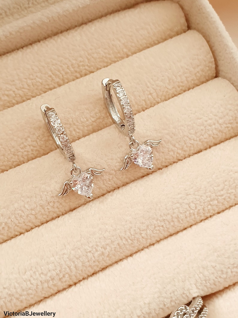 Puede incluir: Un par de pendientes de aro de plata con un dije de coraz&oacute;n colgante. Cada dije de coraz&oacute;n tiene peque&ntilde;as alas a cada lado y est&aacute; adornado con piedras de circonita c&uacute;bica transparentes.