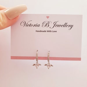 Puede incluir: Aretes de aro de plata con un dije de coraz&oacute;n colgante con alas y piedras de circonita c&uacute;bica transparentes. Los aretes est&aacute;n sobre un fondo blanco con una tarjeta que dice "Victoria B. Jewellery Handmade With Love".