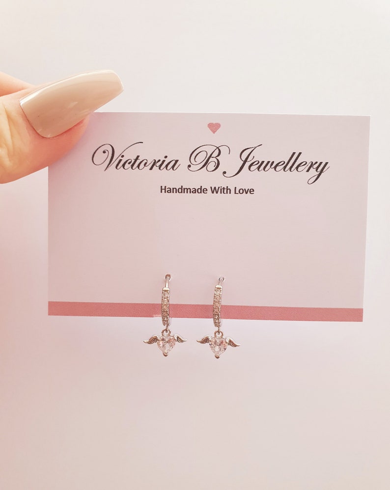 Puede incluir: Aretes de aro de plata con un dije de coraz&oacute;n colgante con alas y piedras de circonita c&uacute;bica transparentes. Los aretes est&aacute;n sobre un fondo blanco con una tarjeta que dice "Victoria B. Jewellery Handmade With Love".