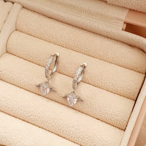 Puede incluir: Un par de pendientes de aro de plata con un peque&ntilde;o dije de coraz&oacute;n con alas que cuelga de cada aro. Los pendientes est&aacute;n en una caja de joyer&iacute;a forrada de terciopelo.