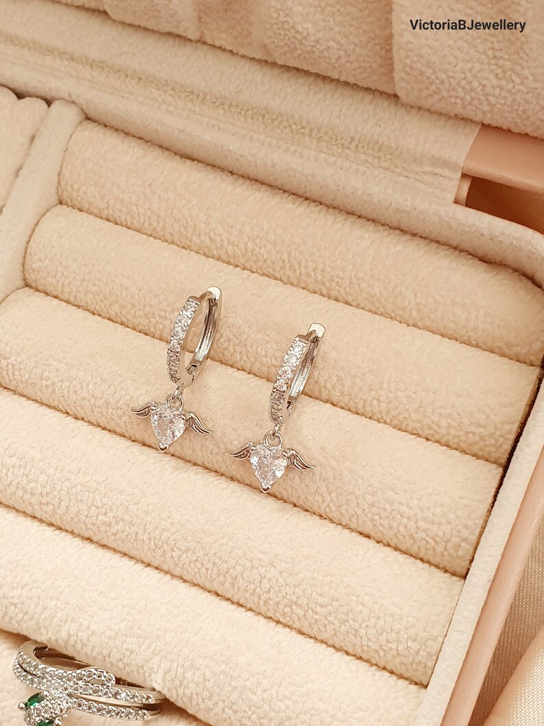 Puede incluir: Un par de pendientes de aro de plata con un peque&ntilde;o dije de coraz&oacute;n con alas que cuelga de cada aro. Los pendientes est&aacute;n en una caja de joyer&iacute;a forrada de terciopelo.