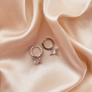 Puede incluir: Un par de pendientes de aro de plata con un peque&ntilde;o dije de &aacute;ngel y una piedra de cristal transparente que cuelga de cada aro.