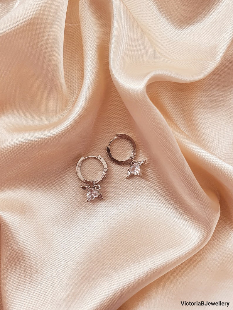 Puede incluir: Un par de pendientes de aro de plata con un peque&ntilde;o dije de &aacute;ngel y una piedra de cristal transparente que cuelga de cada aro.