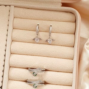 Puede incluir: Un conjunto de pendientes de plata con un dise&ntilde;o de coraz&oacute;n y alas y dos anillos de plata con piedras verdes. Las joyas se muestran en una caja de joyer&iacute;a de terciopelo beige.