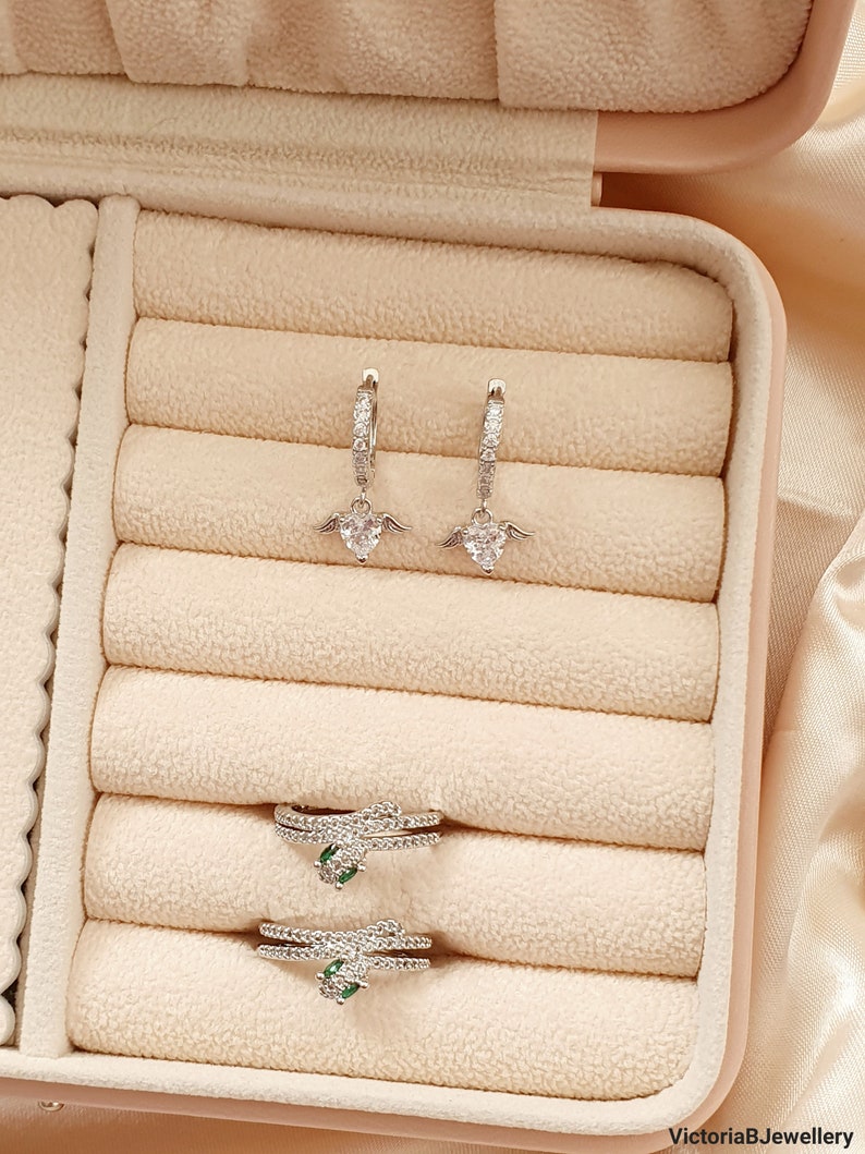 Puede incluir: Un conjunto de pendientes de plata con un dise&ntilde;o de coraz&oacute;n y alas y dos anillos de plata con piedras verdes. Las joyas se muestran en una caja de joyer&iacute;a de terciopelo beige.