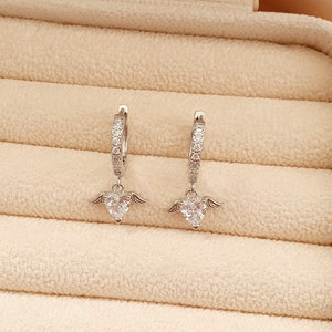 Puede incluir: Un par de pendientes de aro de plata con un colgante de coraz&oacute;n adornado con piedras de circonita c&uacute;bica transparentes. El colgante de coraz&oacute;n tiene peque&ntilde;as alas a cada lado.