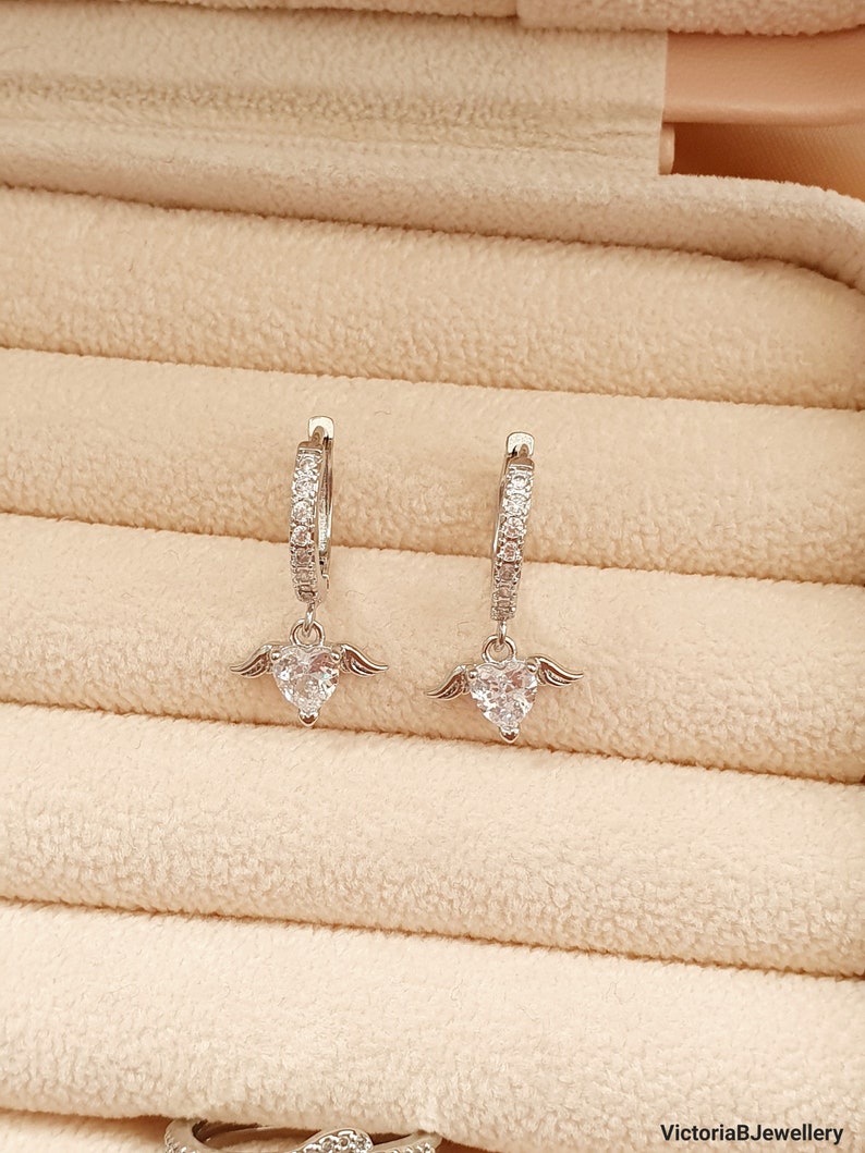 Puede incluir: Un par de pendientes de aro de plata con un colgante de coraz&oacute;n adornado con piedras de circonita c&uacute;bica transparentes. El colgante de coraz&oacute;n tiene peque&ntilde;as alas a cada lado.