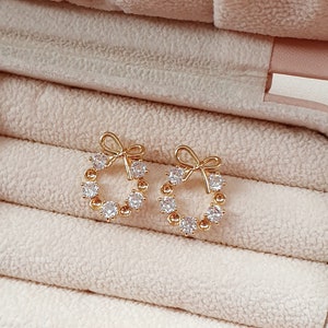 May include: A pair of gold earrings with a circle design and a bow detail. The earrings are set with clear cubic zirconia stones.