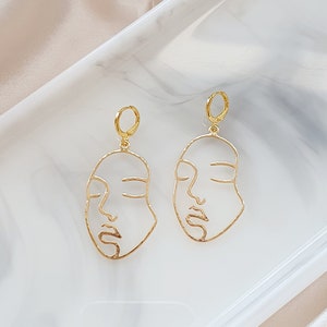 May include: A pair of gold-toned earrings featuring a minimalist line drawing of a face. The earrings are dangling and have a hoop closure.