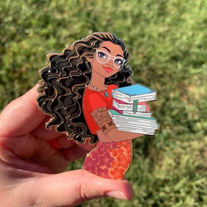 Op de afbeelding: Een gouden emaille pin met een cartoon vrouw met lang krullend bruin haar, die rode kleding, een bril en een ketting draagt. Ze houdt een stapel kleurrijke boeken vast.