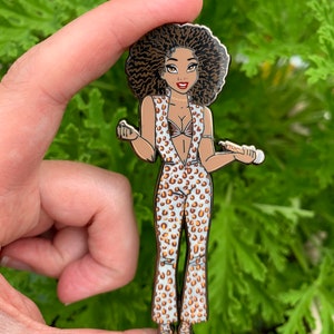 May include: A brown and white enamel pin featuring a cartoon woman with an afro hairstyle wearing a leopard print jumpsuit and holding a microphone.