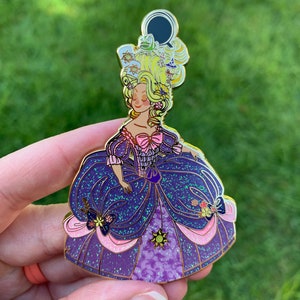Pu&ograve; includere: Uno spillo in smalto dorato con una principessa dei cartoni animati con i capelli biondi e un vestito viola e rosa. La principessa indossa una corona con una rana e un sole sopra. L'abito &egrave; ricoperto di glitter.