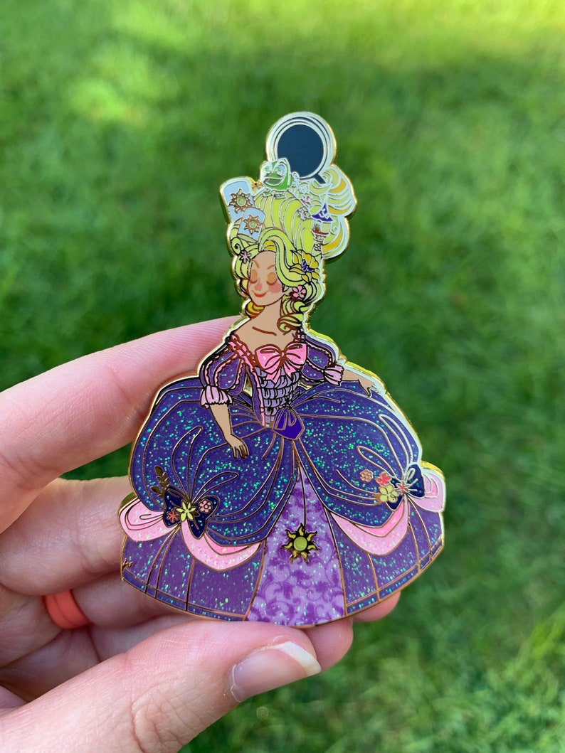 Pu&ograve; includere: Uno spillo in smalto dorato con una principessa dei cartoni animati con i capelli biondi e un vestito viola e rosa. La principessa indossa una corona con una rana e un sole sopra. L'abito &egrave; ricoperto di glitter.