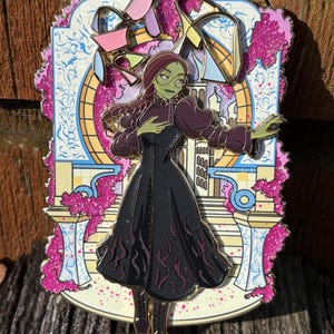 Op de afbeelding: Een kleurrijke emaille pin met een gestileerde illustratie van een groenhuidige vrouw in een zwarte jurk met een paarse en roze achtergrond. De pin heeft een goudkleurige omtrek en toont een scène met architecturale elementen.