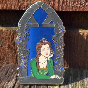 Puede incluir: Pin metálico que representa a una princesa en una ventana. La princesa tiene el pelo rojo, un vestido verde y una tiara. El marco de la ventana es gris y el fondo es azul. Enredaderas con flores azules decoran el marco.