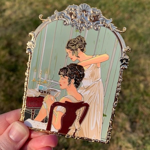 May include: Gold-toned enamel pin depicting two women in a vintage setting. One woman is seated and the other is standing behind her, adjusting her hair. The pin has a detailed ornate frame.