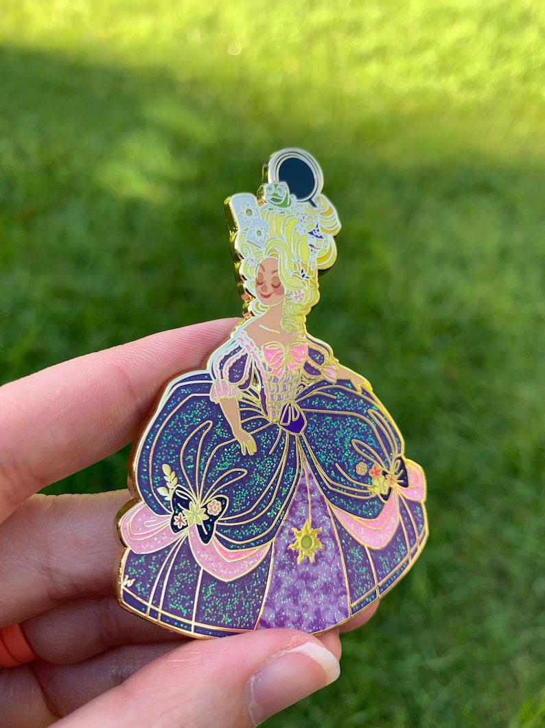Pu&ograve; includere: Uno spillo in smalto dorato con una rappresentazione di un cartone animato di una principessa con un abito viola e rosa con accenti di glitter. La principessa ha i capelli biondi e indossa una corona con un design di sole e luna.