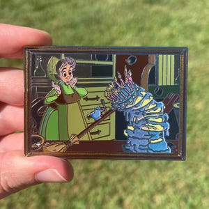 May include: A rectangular enamel pin featuring a cartoon scene with a woman in a green dress and a tall, blue cake with lit candles. The pin has a gold border and depicts a kitchen setting.