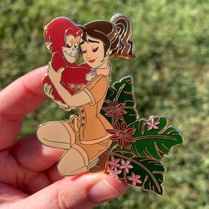 Peut inclure: Une &eacute;pingle en &eacute;mail dor&eacute;e repr&eacute;sentant une femme en tenue de safari kaki tenant un singe rouge. La femme sourit et le singe la regarde. L'&eacute;pingle est sur un fond de feuilles vertes avec des fleurs roses.