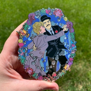 May include: A blue and white enamel pin featuring a couple dancing underwater. The pin is surrounded by colorful sea creatures and bubbles. The couple is dressed in formal attire, with the man wearing a black suit and the woman wearing a purple dress.