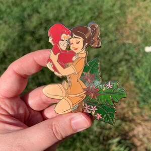 Peut inclure: Une &eacute;pingle en &eacute;mail dor&eacute;e repr&eacute;sentant une femme aux cheveux bruns tenant un orang-outan rouge. La femme porte une tenue de safari beige et est entour&eacute;e de feuilles vertes et de fleurs roses.