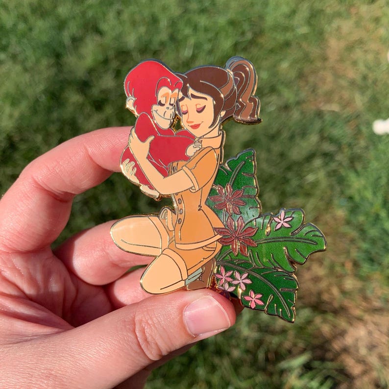 Peut inclure: Une &eacute;pingle en &eacute;mail dor&eacute;e repr&eacute;sentant une femme aux cheveux bruns tenant un orang-outan rouge. La femme porte une tenue de safari beige et est entour&eacute;e de feuilles vertes et de fleurs roses.